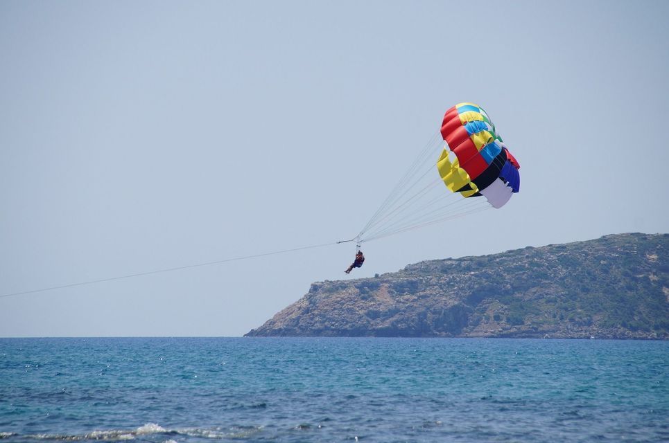 Una persona che fa parasailing con un paracadute colorato scivola sul mare blu con un promontorio roccioso sullo sfondo.