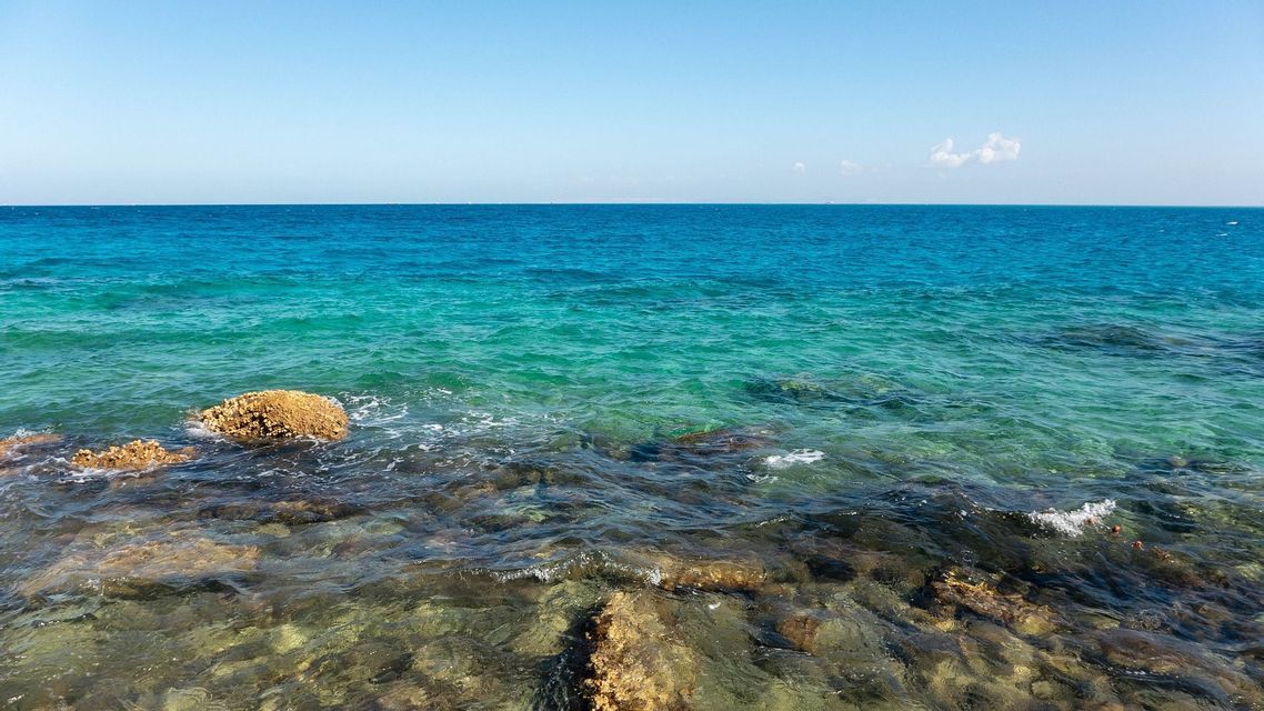 Rocce visibili attraverso acqua limpida e poco profonda al bordo di un mare calmo e turchese sotto un cielo azzurro brillante.