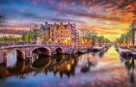 Un ponte ad arco su un canale con case tradizionali e barche riflesse nell'acqua durante un tramonto colorato.