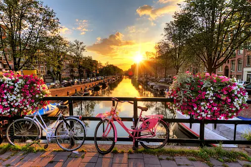 Due biciclette parcheggiate su un ponte adornato con cesti di fiori, con vista su un canale cittadino al tramonto.