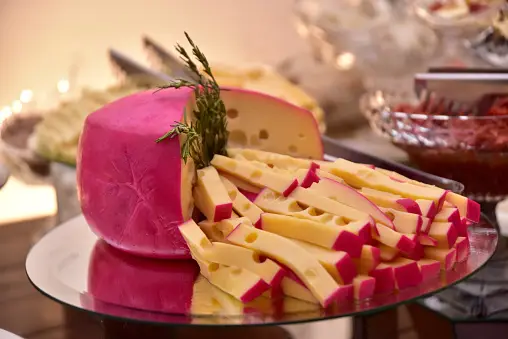 Una forma di formaggio tipo svizzero con crosta di cera rosa, affettato e disposto su un piatto a specchio con guarnizione di rosmarino.