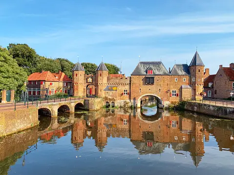 Un casello in mattoni medievale e un ponte si riflettono nell'acqua calma di un canale sotto un cielo azzurro chiaro.