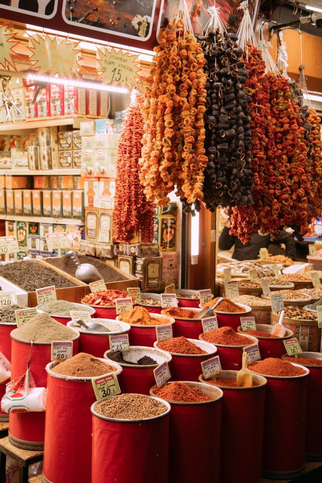 Una bancarella del mercato espone varie spezie e cereali in grandi contenitori rossi, con filze di peperoni secchi e verdure appese in alto.