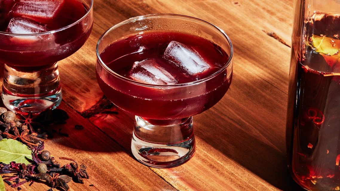 Un bicchiere di tè all'ibisco rosso intenso con cubetti di ghiaccio su un tavolo di legno, accanto a una brocca e spezie essiccate.