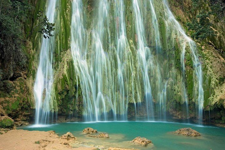 Una larga cascata scende da una scogliera coperta di muschio in una pozza d'acqua turchese con una riva sabbiosa.