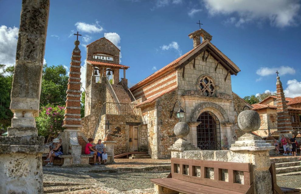 Una chiesa rustica in pietra con campanile e un obelisco in una piazza soleggiata con persone che riposano sulle panchine.