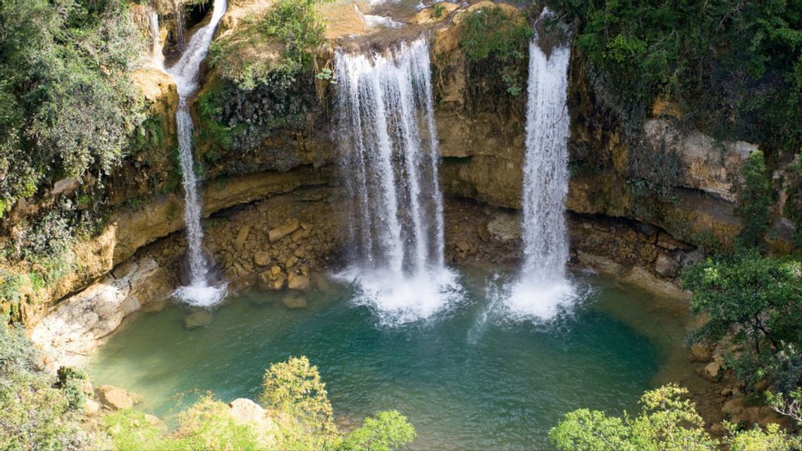 Diverse cascate scendono da una scogliera rocciosa in una piscina turchese circondata da una lussureggiante vegetazione verde.