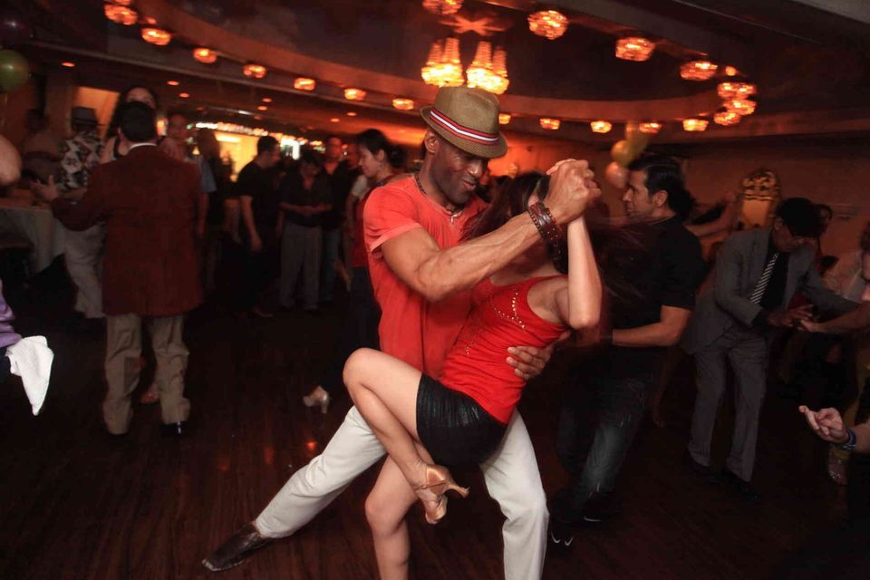 Un uomo con un fedora e una donna con un top rosso ballano salsa insieme su una pista da ballo affollata e poco illuminata.