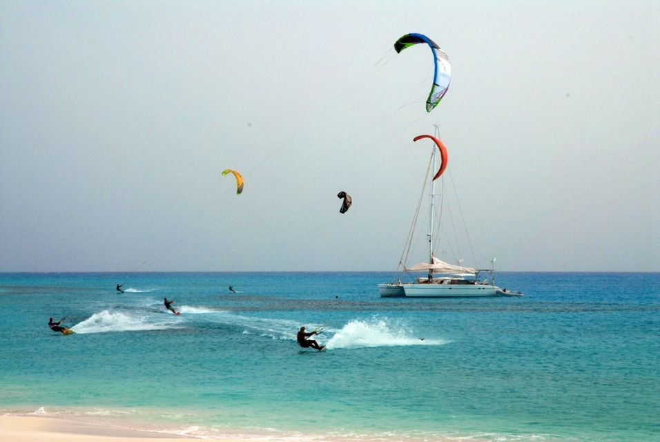 Diversi kitesurfer scivolano sull'acqua turchese con un catamarano a vela bianco ancorato sullo sfondo