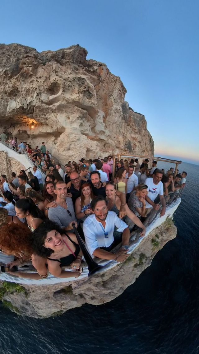 Un groupe WeRoad prend un selfie grand-angle sur une terrasse de bar bondée, à flanc de falaise, au-dessus de la mer.
