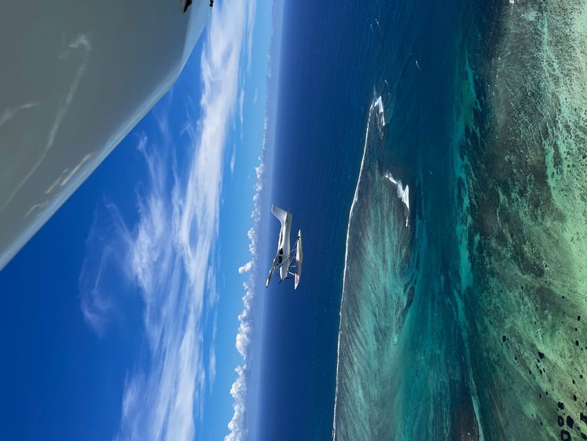 Veduta aerea di un trimarano a vela bianco su acqua turchese vicino a una grande barriera corallina sotto un cielo blu con nuvole.