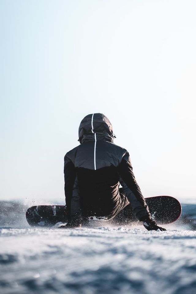 Eine Person in schwarzer Kapuzenjacke sitzt im Schnee auf einem Snowboard, von hinten vor hellem Himmel gesehen.