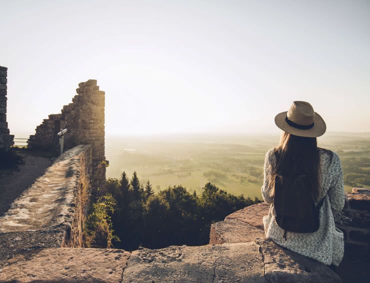 Eine Frau mit Hut und Rucksack sitzt am Rande einer Steinruine und blickt auf ein sonnendurchflutetes Tal und einen Wald.