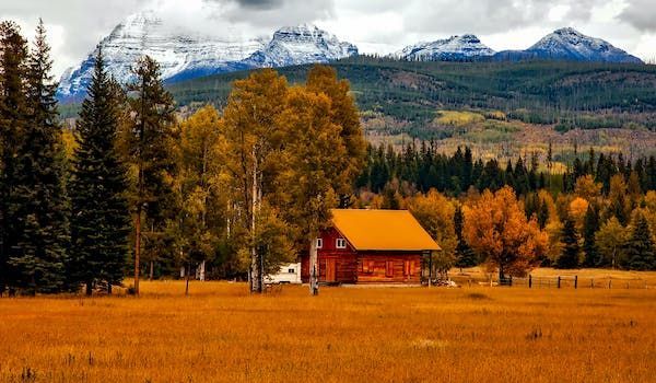Una baita di legno con un tetto arancione si trova in un campo dorato, con alberi autunnali e montagne innevate sullo sfondo.