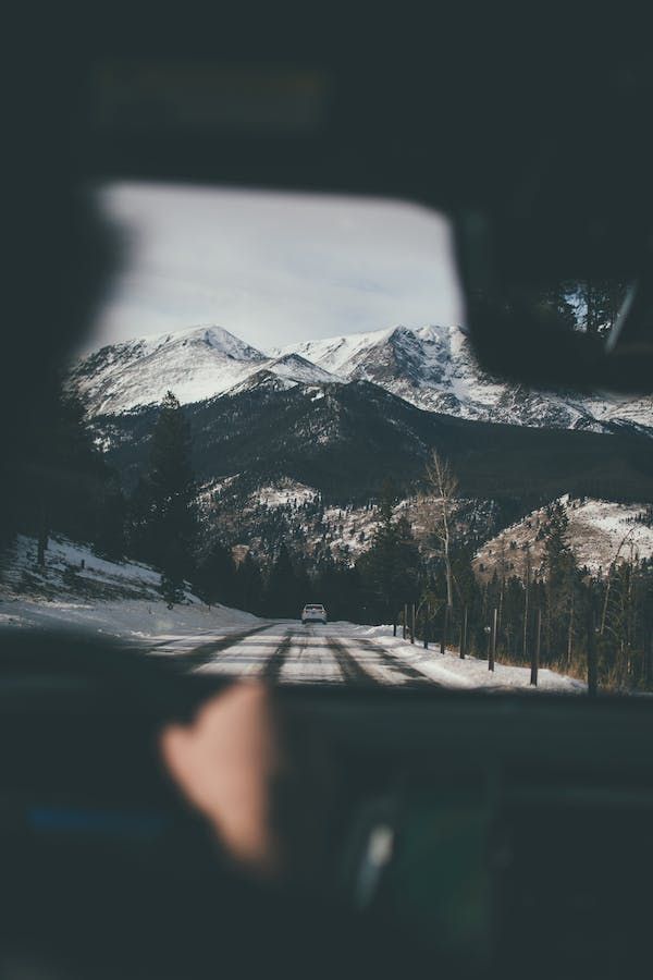 Una vista dall'interno di un veicolo, con un'auto bianca che avanza su una strada innevata verso imponenti montagne coperte di neve.