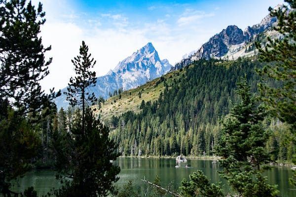 Una vetta montuosa frastagliata si erge su una fitta pineta e un lago sereno, incorniciata da alberi sempreverdi sotto un cielo azzurro.