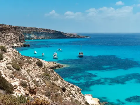 Una vista dall'alto di diverse barche bianche ancorate in una cala rocciosa con acqua cristallina turchese e blu profondo sotto un cielo azzurro.