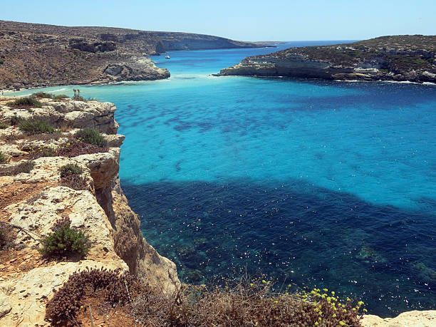 Vista da una scogliera rocciosa su una baia assolata, con acque turchesi e blu profondo.