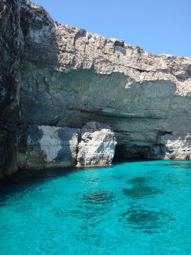 Una scogliera rocciosa con grotte marine alla base incontra acque turchesi brillanti sotto un cielo azzurro limpido.