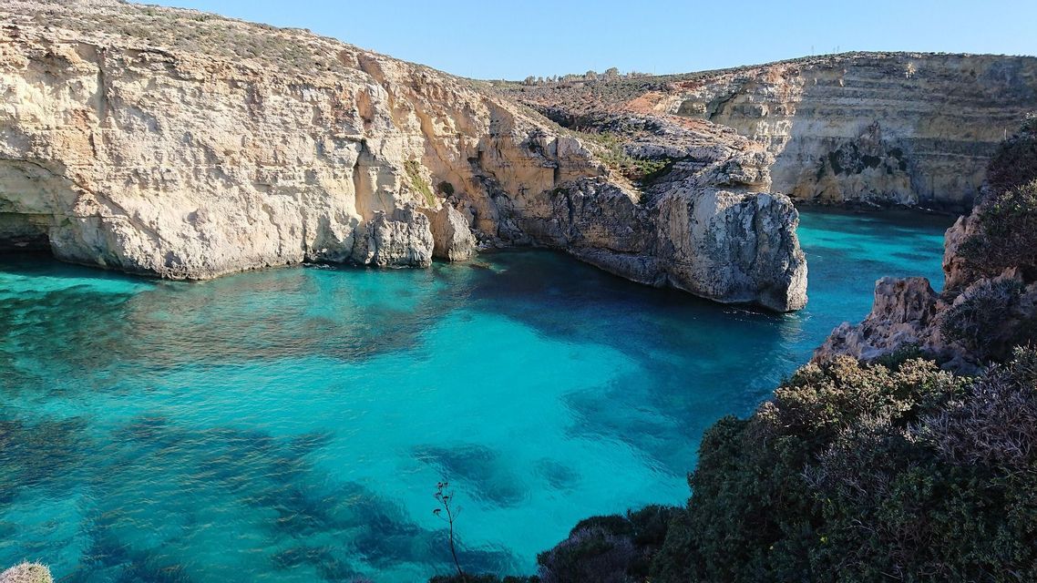 Una caletta con acqua cristallina e turchese è circondata da alte scogliere rocciose sotto un cielo azzurro brillante.