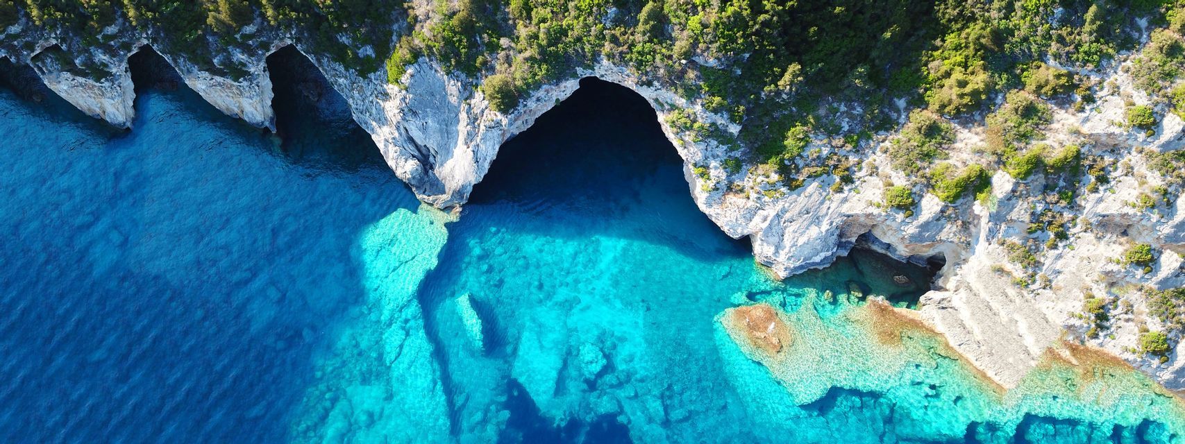 An aerial top-down view of sea caves in a white rocky cliff with green vegetation, surrounded by turquoise and deep blue water.