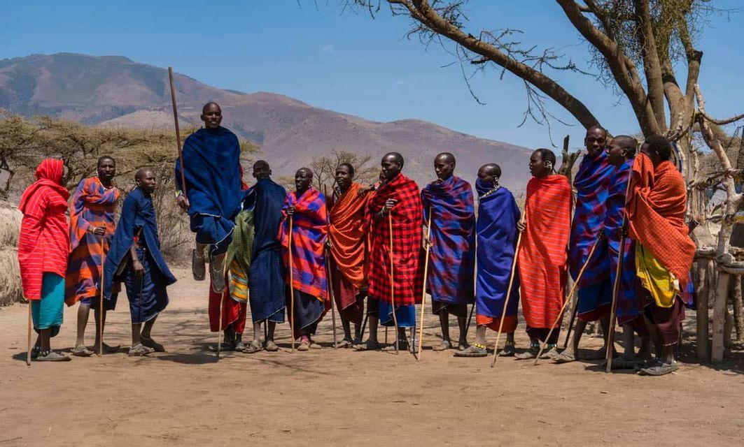 Un gruppo di uomini Maasai in tuniche tradizionali colorate si allinea, con un uomo che salta, di fronte a un paesaggio montano.