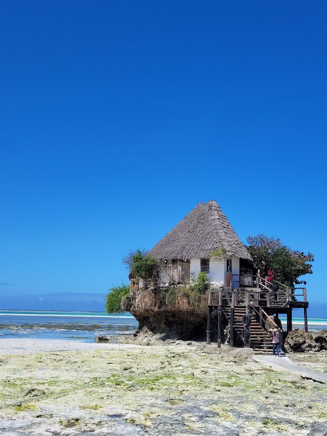Ein reetgedecktes Gebäude thront auf einem großen Felsen an einem Strand bei Ebbe unter klarem blauem Himmel.