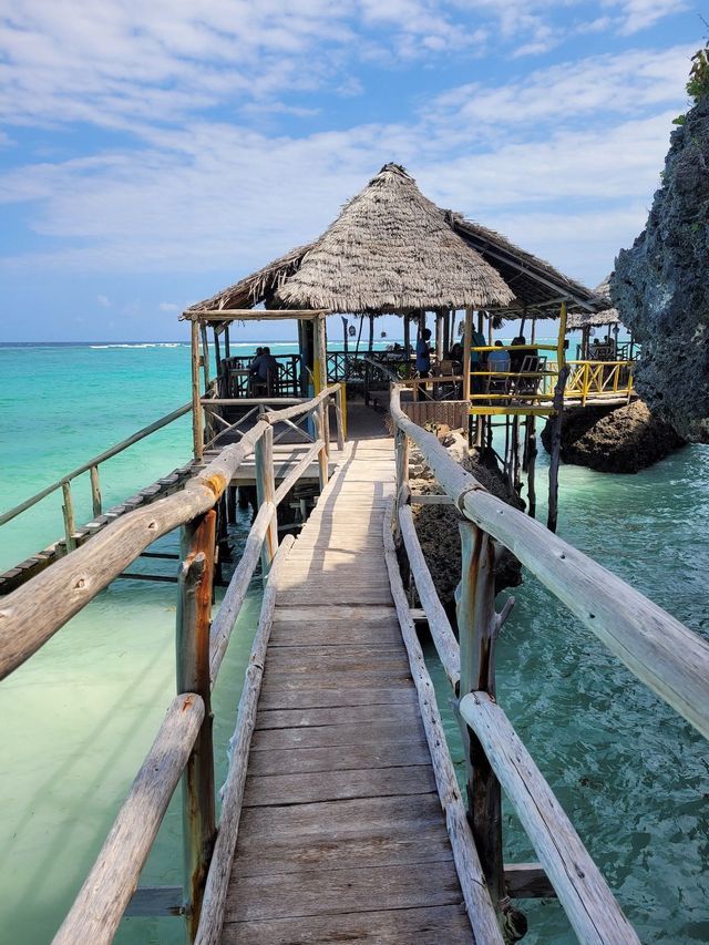 Ein Holzsteg führt zu einem Reetdachrestaurant, das auf Stelzen über türkisfarbenem Wasser unter einem teils bewölkten Himmel steht.