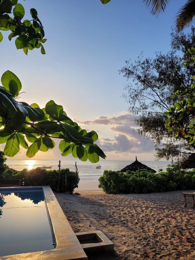 Die Sonne geht über dem Ozean auf, gesehen von einem Sandstrand mit einem Poolbereich und üppiger tropischer Vegetation.