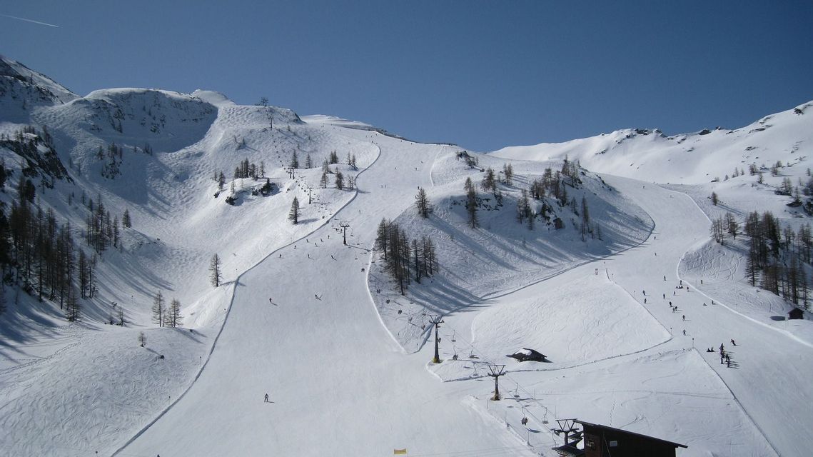 Un'ampia vista di una stazione sciistica in una giornata di sole, con numerose persone che sciano su piste battute tra alberi sparsi e seggiovie.
