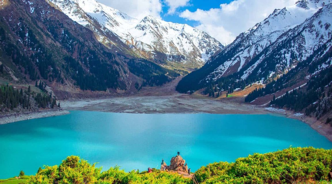 Una vista panoramica di un lago alpino turchese in una valle, con massicce montagne innevate sullo sfondo.