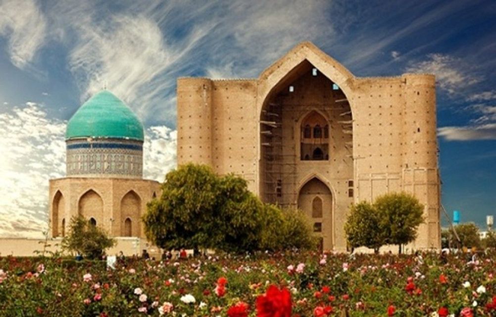 A sand-colored mausoleum with a large archway and a turquoise dome, viewed from behind a colorful garden of roses.