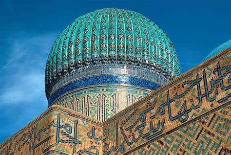 An ornate, turquoise-tiled dome and walls with intricate calligraphy sit against a clear blue sky.