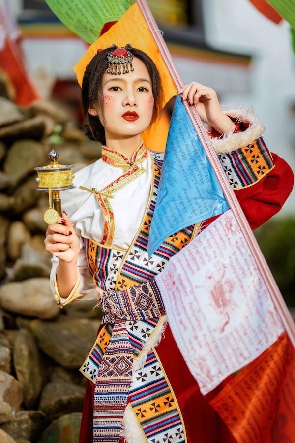 Una donna in abito tradizionale tibetano tiene una ruota di preghiera dorata e una serie di bandiere di preghiera colorate, guardando la macchina fotografica.