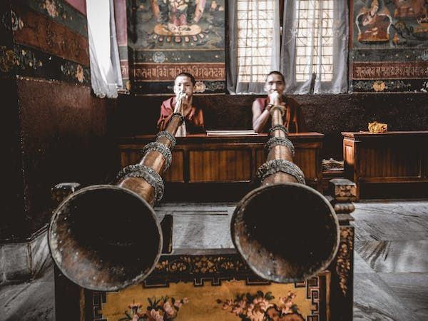 Due monaci in tuniche rosse siedono dietro due grandi, ornati corni tradizionali all'interno di un monastero con pareti dipinte.