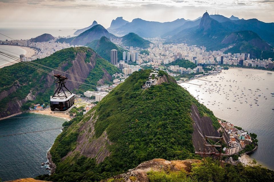 A cable car travels over a lush green mountain, with a panoramic view of a coastal city, a bay with boats, and distant mountains.