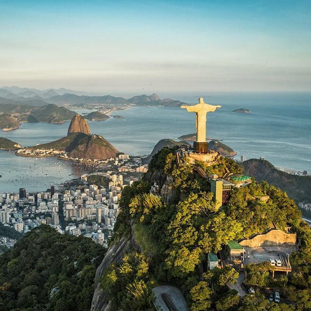 An aerial view of the Christ the Redeemer statue on a mountain overlooking a coastal city, a bay, and Sugarloaf Mountain under a clear sky.