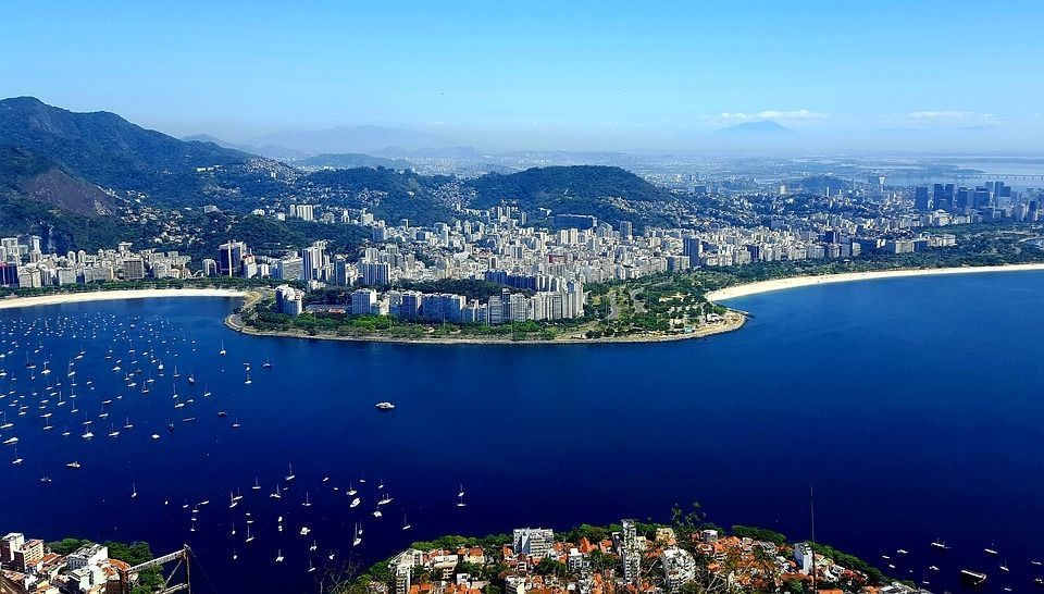 An aerial view of a coastal city along a bay filled with sailboats, nestled against green hills under a clear blue sky.