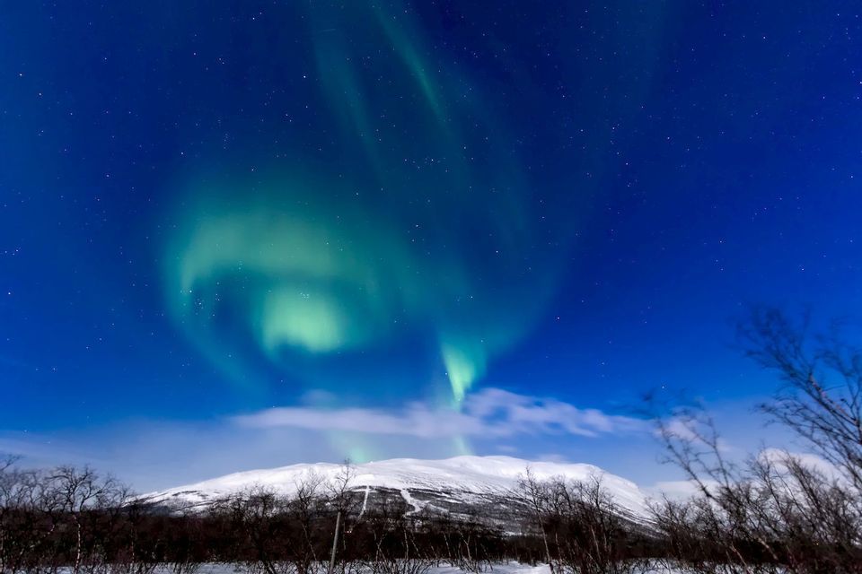 La aurora boreal verde se arremolina en un cielo nocturno estrellado sobre un paisaje montañoso cubierto de nieve.