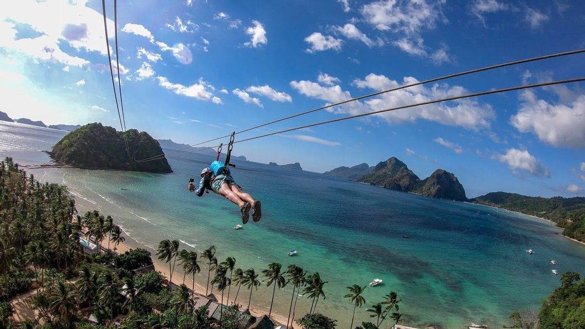 A person on a zip-line glides over a tropical beach with palm trees, turquoise water, and distant islands.