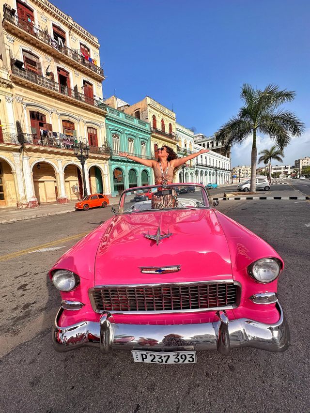 Eine Frau steht mit ausgestreckten Armen in einem knallrosa Vintage-Cabrio auf einer Stadtstraße, gesäumt von bunten Gebäuden und einer Palme.