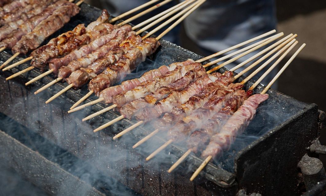 Fila di spiedini di carne alla griglia su un barbecue lungo e stretto con fumo che sale dalle braci.