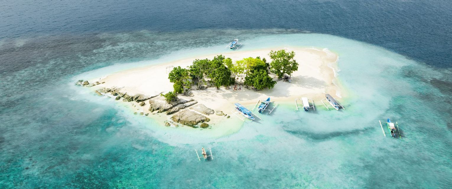 Vue aérienne d'une petite île tropicale avec une plage de sable blanc, des arbres verts et plusieurs bateaux dans l'eau turquoise.