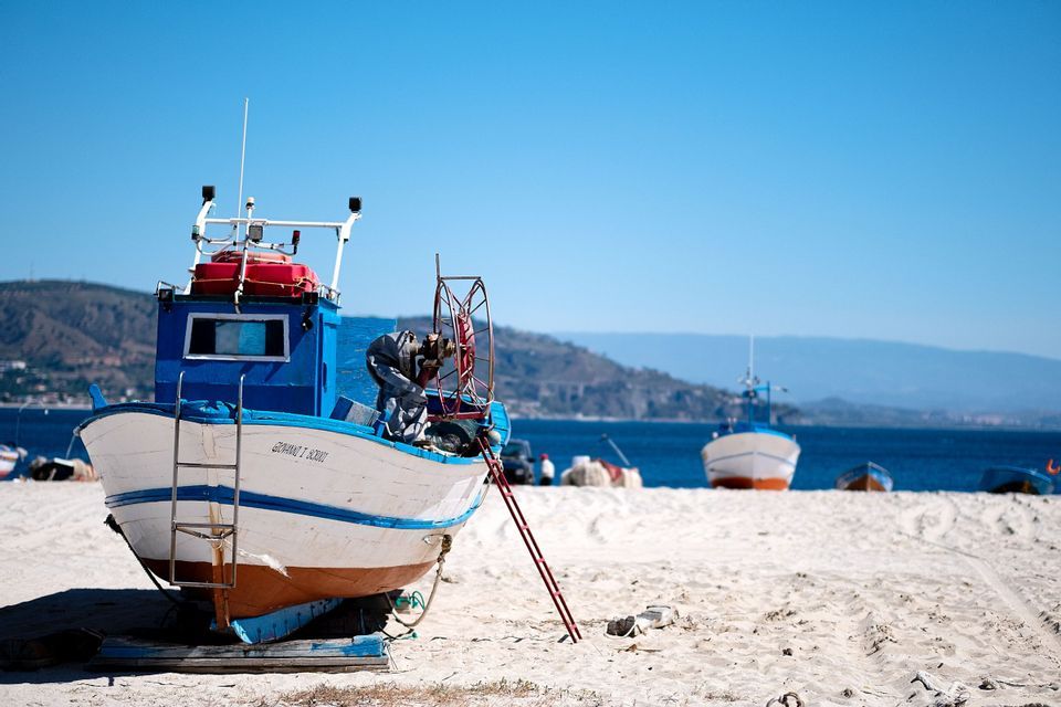 Ein blau-weißes Fischerboot liegt an einem Sandstrand mit dem Meer und fernen Hügeln im Hintergrund unter einem klaren Himmel.