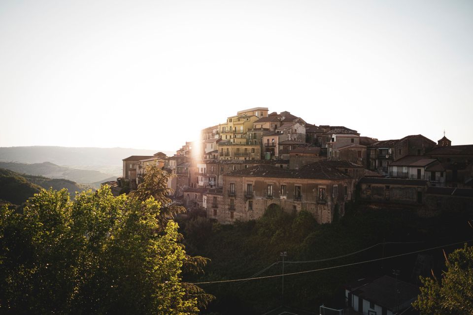 Ein Dorf mit alten Häusern, erbaut an einem grünen Hang, wobei die aufgehende Sonne einen hellen Schein hinter den Gebäuden erzeugt.