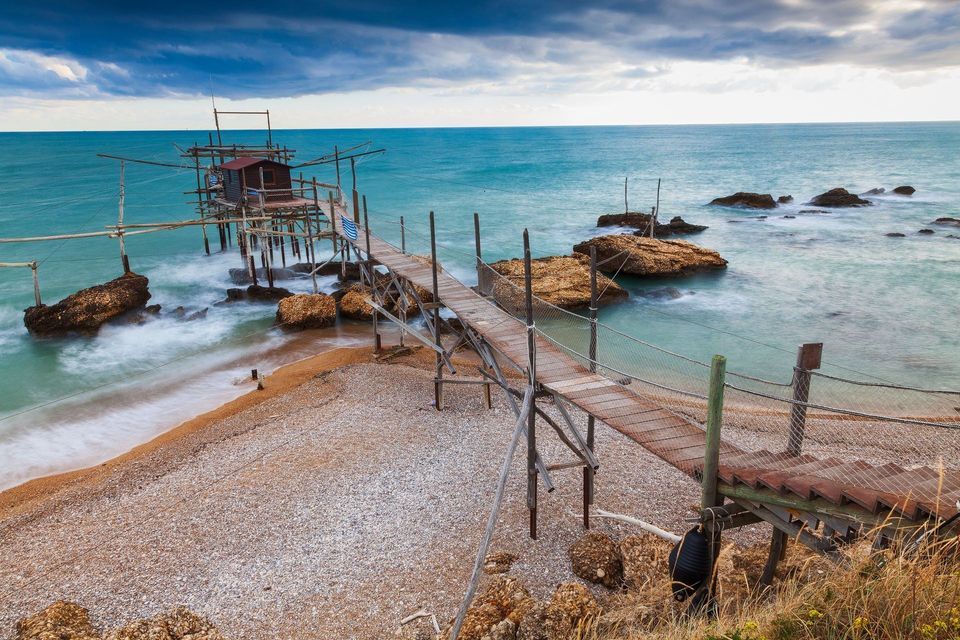 Un lungo molo di legno conduce da una spiaggia di ciottoli a una capanna di pescatori su palafitte nel mare turchese sotto un cielo nuvoloso.