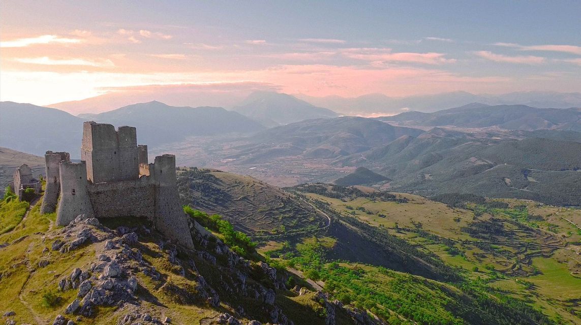 Una rovina di castello in pietra si erge su una collina rocciosa che domina una vasta valle di montagna verde sotto un cielo dai toni rosati.