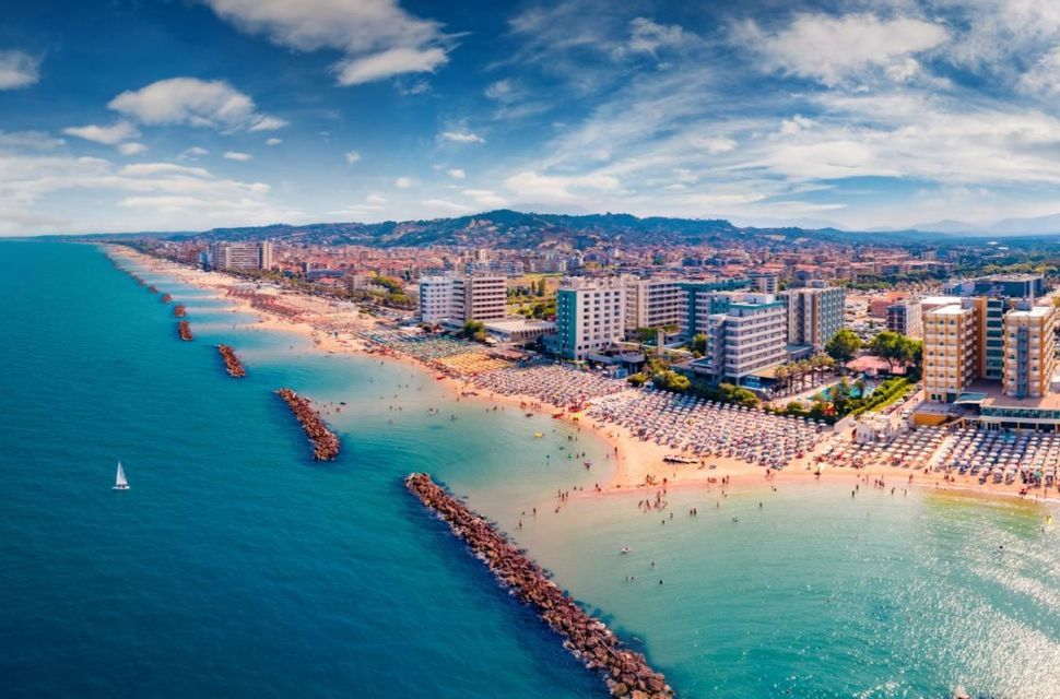 Vista aerea di una lunga spiaggia sabbiosa affollata di ombrelloni e persone, accanto a una città con colline sullo sfondo.