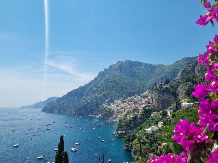 Un pueblo costero construido en una ladera empinada y verde con vistas a una bahía azul llena de barcos, con flores rosas en primer plano.