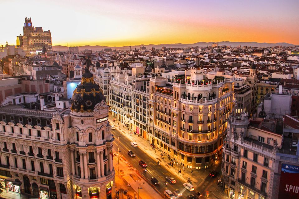 Vista aerea di una fitta città con edifici ornati e strade illuminate al tramonto, con montagne lontane all'orizzonte.